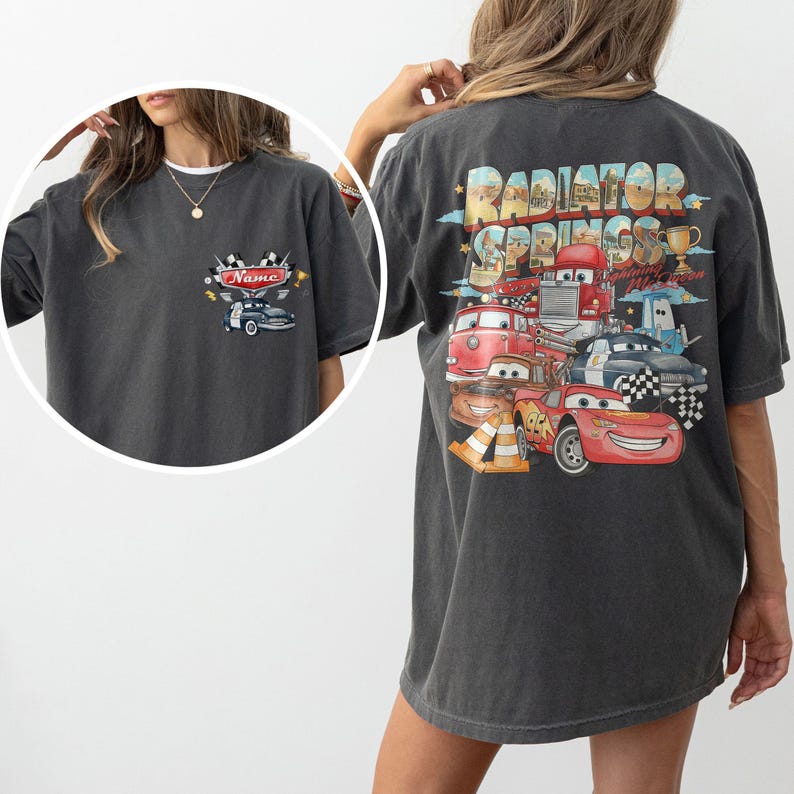 May include: A charcoal gray t-shirt featuring a small graphic on the front left chest with a car and the text Name. The back displays a large, colorful graphic of Radiator Springs characters including Lightning McQueen, with the text Radiator Springs.