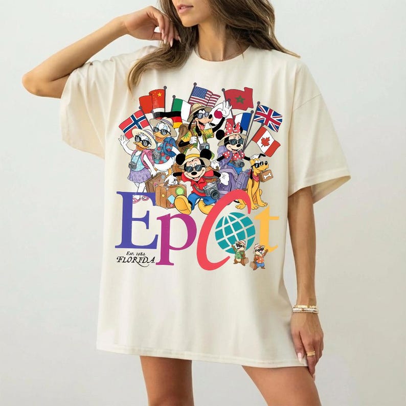 K&ouml;nnte beinhalten: Cremefarbenes T-Shirt mit einem farbenfrohen Aufdruck von Disney-Figuren, die Flaggen aus aller Welt halten. Das Wort "EPCOT" ist in gro&szlig;en Buchstaben dargestellt, darunter "Est. 1982 FLORIDA". Das Shirt hat kurze &Auml;rmel und eine lockere Passform.