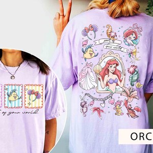 May include: Orchid-colored t-shirt featuring a colorful graphic of Ariel from The Little Mermaid, with the text "Part of your world." The design includes various characters and elements from the movie. The shirt is a comfortable, casual style.
