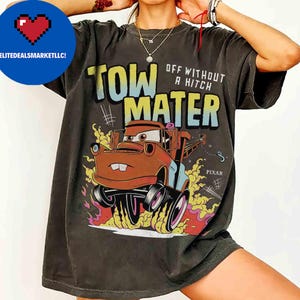 May include: Dark grey t-shirt with a cartoon image of Tow Mater from the film Cars. The graphic includes the text "TOW MATER" and "OFF WITHOUT A HITCH" in colourful lettering, with the Pixar logo. The shirt has a vintage, worn appearance.