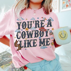 May include: A light pink t-shirt featuring the dark blue text "YOU'RE A COWBOY LIKE ME" surrounded by colourful graphics including a rocket, a cowboy hat, a sheriff badge, alphabet blocks, a ball, clouds, stars, and a pair of brown cowboy boots.