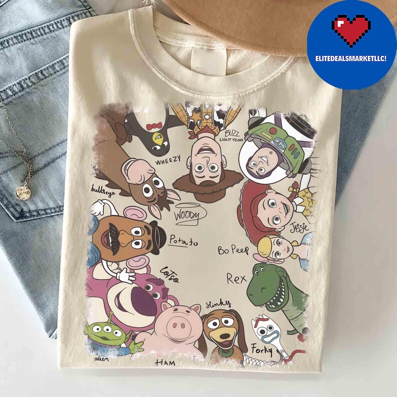 May include: Cream-colored t-shirt featuring a colorful graphic of Toy Story characters, including Woody, Buzz Lightyear, and Jessie. The design includes character names and is surrounded by a blue circle with a heart and the text "ELITEDEALSMARKETLLC!".