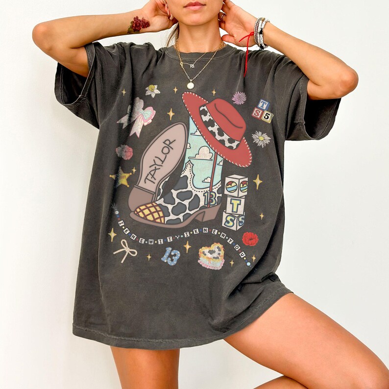 Puede incluir: Una camiseta gris carb&oacute;n de manga corta con un estampado gr&aacute;fico de una bota vaquera con estampado de vaca, un sombrero vaquero rojo y el nombre Taylor. Los gr&aacute;ficos adicionales incluyen bloques con las letras TS, el n&uacute;mero 13, estrellas, lazos y flores.