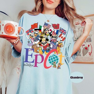 K&ouml;nnte beinhalten: Hellblaues T-Shirt mit einer farbenfrohen Grafik mit Disney-Figuren, Flaggen und dem Text "Epcot Florida Est. 1982". Das Shirt hat kurze &Auml;rmel und eine lockere Passform. Eine wei&szlig;e Tasse mit einem Sonnendesign wird im Bild gehalten.