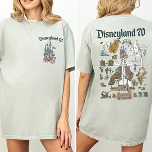 Puede incluir: Una camiseta de color verde salvia claro con un peque&ntilde;o logotipo de Disneyland 70 y un gr&aacute;fico de castillo en el pecho izquierdo. La parte posterior muestra una ilustraci&oacute;n grande y detallada de las atracciones de Disneyland con el texto Disneyland 70.