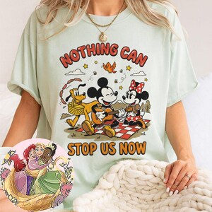 May include: A light green t-shirt featuring a graphic of Mickey Mouse playing a guitar, Minnie Mouse, and Pluto on a picnic blanket. The text reads NOTHING CAN STOP US NOW. A circular inset shows an illustration of three animated characters hugging.