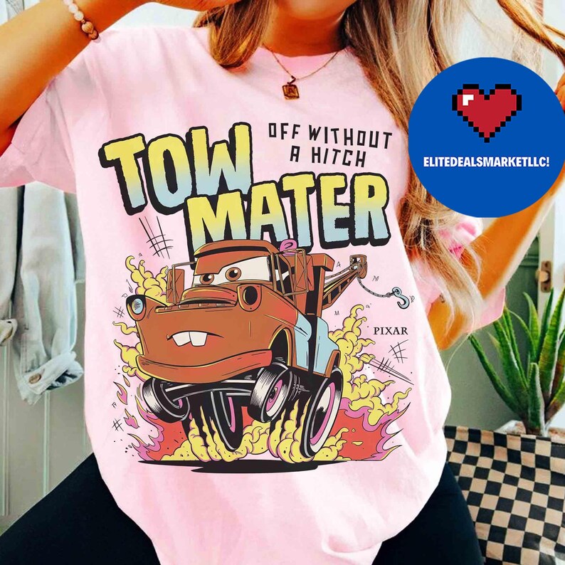 May include: Pink t-shirt with a cartoon image of Tow Mater from the film Cars. The graphic includes the text "TOW MATER" in yellow and blue, with "OFF WITHOUT A HITCH" above the character. The design is surrounded by flames.