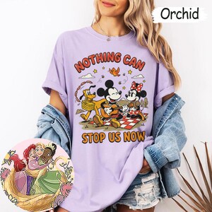 May include: An orchid-colored t-shirt featuring a graphic of Mickey Mouse playing a guitar, Minnie Mouse, and Pluto on a picnic. The text reads NOTHING CAN STOP US NOW. A circular inset shows an illustration of three animated characters embracing.