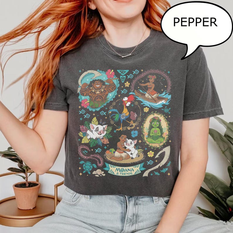 May include: Dark gray t-shirt featuring a colorful graphic of characters from the animated film Moana. The design includes images of Maui, Moana, Hei Hei, Pua, and other characters. The word "Pepper" is in a speech bubble.