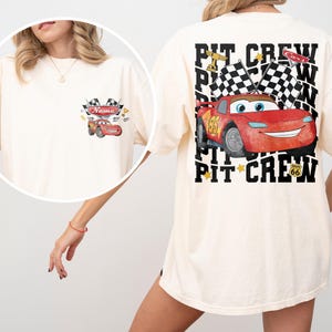 May include: An off-white t-shirt featuring a red race car graphic on the front left chest and a large back print. The back design displays a red race car, checkered flags, and the text PIT CREW with a Route 66 sign. The front graphic includes the word Name.