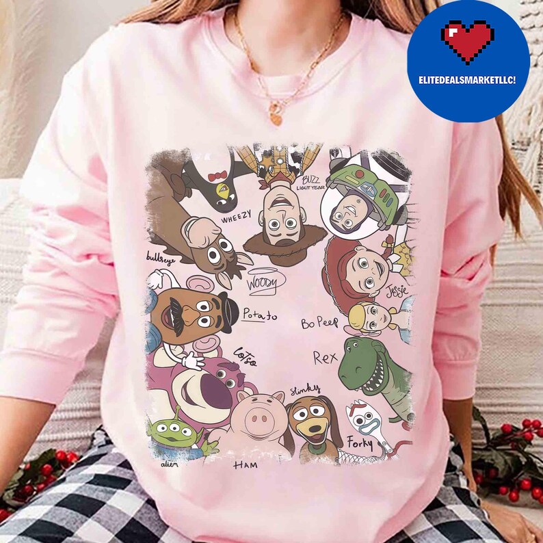 May include: A light pink sweatshirt featuring a colorful graphic of Toy Story characters. The design includes Woody, Buzz Lightyear, Jessie, and others, with character names written below. The sweatshirt has long sleeves and a crew neck.
