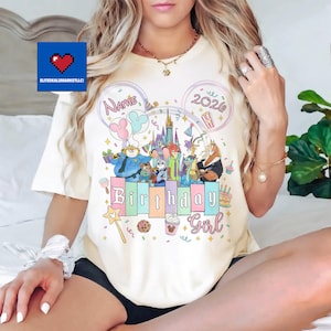 May include: Cream-colored t-shirt featuring a colorful graphic with cartoon characters, a castle, balloons, and the words "Birthday Girl" and "2020". The design includes a space for a name. The shirt has a round neckline.