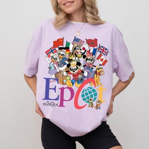 K&ouml;nnte beinhalten: Ein helllila T-Shirt mit einem farbenfrohen Aufdruck von Zeichentrickfiguren, die Flaggen aus aller Welt halten. Das Wort "EPCOT" ist prominent dargestellt, zusammen mit dem Text "Est. 1982 Florida". Das Design beinhaltet einen Globus und Reiseelemente.