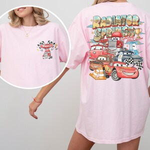 May include: A light pink t-shirt featuring a small graphic on the front left chest with a red truck, checkered flags, and the text Name. The back displays a large Radiator Springs graphic with Lightning McQueen, various cars, and a gold trophy.