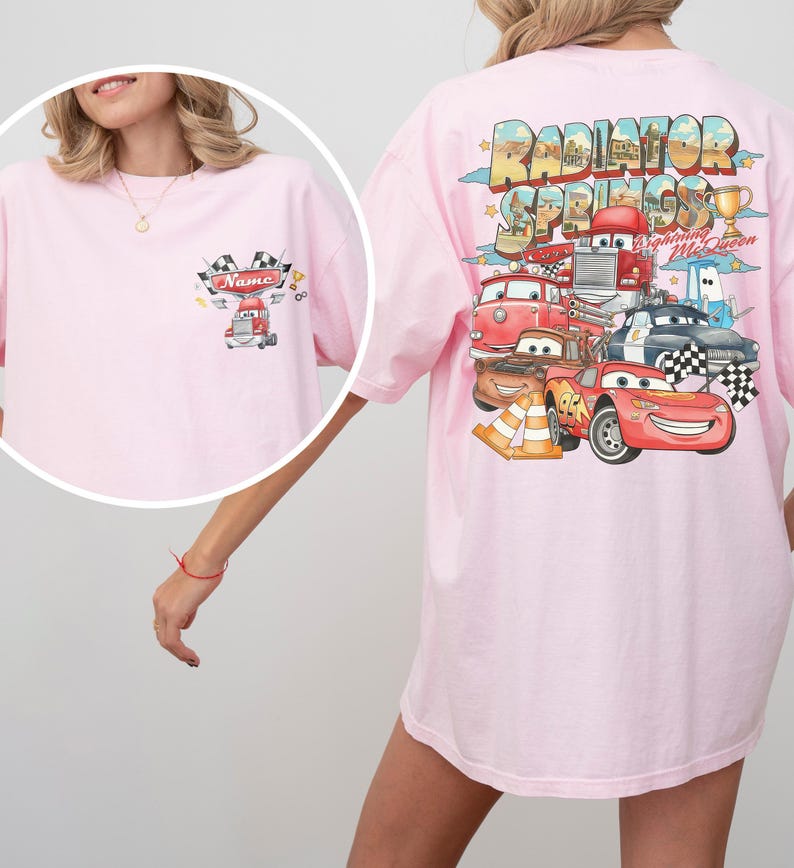 May include: A light pink t-shirt featuring a small graphic on the front left chest with a red truck, checkered flags, and the text Name. The back displays a large Radiator Springs graphic with Lightning McQueen, various cars, and a gold trophy.