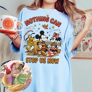 May include: A light blue t-shirt featuring a graphic of Mickey Mouse playing a guitar, Minnie Mouse, and Pluto on a picnic blanket. The text reads "NOTHING CAN STOP US NOW". A circular inset shows an illustration of three animated characters hugging.