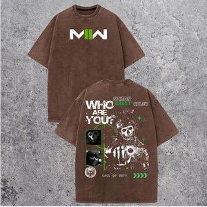 May include: Brown t-shirt with the letters "MW" in white and green on the front. The back of the shirt features a graphic of a skull and the text "WHO ARE YOU?" and "CALL OF DUTY".