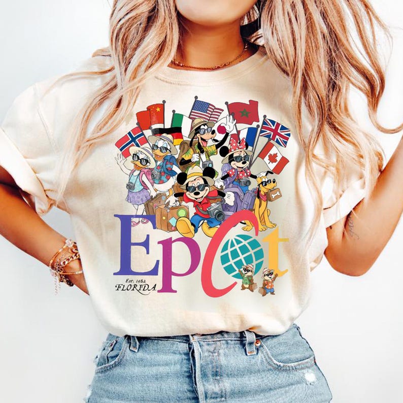 K&ouml;nnte beinhalten: Cremefarbenes T-Shirt mit einer farbenfrohen Grafik von Disney-Figuren, darunter Mickey und Minnie Mouse, Goofy, Donald und Daisy Duck sowie Pluto, die Flaggen aus aller Welt halten. Das Wort "Epcot" ist in Lila und Rot dargestellt, darunter "Est. 1982 Florida".