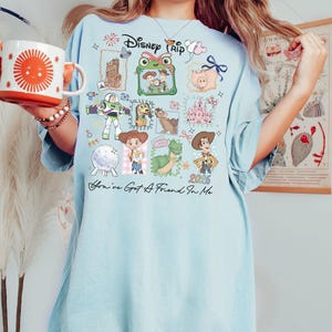 May include: A light blue t-shirt featuring a graphic print with the text Disney Trip and You've Got A Friend In Me. The design includes illustrations of various animated characters, a castle, and the year 2026 arranged in a collage style.