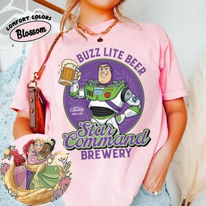 Może przedstawiać: Jasnor&oacute;żowy t-shirt z grafiką przedstawiającą strażnika kosmosu trzymającego kufel piwa wewnątrz fioletowego koła. Napis głosi BUZZ LITE BEER STAR COMMAND BREWERY. Okrągła wstawka przedstawia trzy animowane postacie z długimi włosami, kt&oacute;re się przytulają.