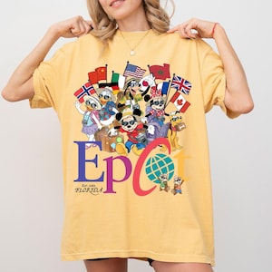 K&ouml;nnte beinhalten: Senfgelbes T-Shirt mit einem farbenfrohen Grafikdesign von Disney-Figuren, die Flaggen aus aller Welt halten. Das Wort "Epcot" ist gro&szlig; geschrieben, darunter steht "Est. 1982 Florida". Das T-Shirt hat eine lockere Passform.