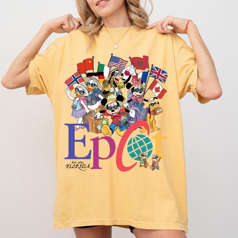 K&ouml;nnte beinhalten: Senfgelbes T-Shirt mit einem farbenfrohen Grafikdesign von Disney-Figuren, die Flaggen aus aller Welt halten. Das Wort "Epcot" ist gro&szlig; geschrieben, darunter steht "Est. 1982 Florida". Das T-Shirt hat eine lockere Passform.