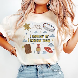 May include: A cream-colored t-shirt featuring a graphic print with the text "I KNEW IT I KNEW YOU", "infinity and beyond", and "YOU'VE GOT A FRIEND IN ME". The design includes a gold sheriff badge, a beaded bracelet, cowboy boots, blocks, and a hat.