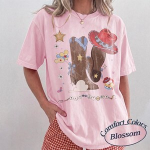 May include: A light pink short-sleeve t-shirt featuring a graphic print of brown cowboy boots, a red cowboy hat, a sheriff star, balloons, and flowers. The text TAY STORY is printed in a circular pattern. The shirt has a crew neckline.
