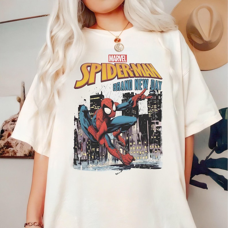 May include: Off-white t-shirt with a Spider-Man graphic, set against a city backdrop. The design includes the Marvel logo and the text "SPIDER-MAN BRAND NEW DAY" in yellow and blue.