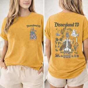 Puede incluir: Una camiseta de color amarillo mostaza con un peque&ntilde;o gr&aacute;fico de un castillo y el texto Disneyland 70 en el pecho izquierdo. La parte trasera muestra una gran ilustraci&oacute;n detallada de varias atracciones y el texto Disneyland 70.