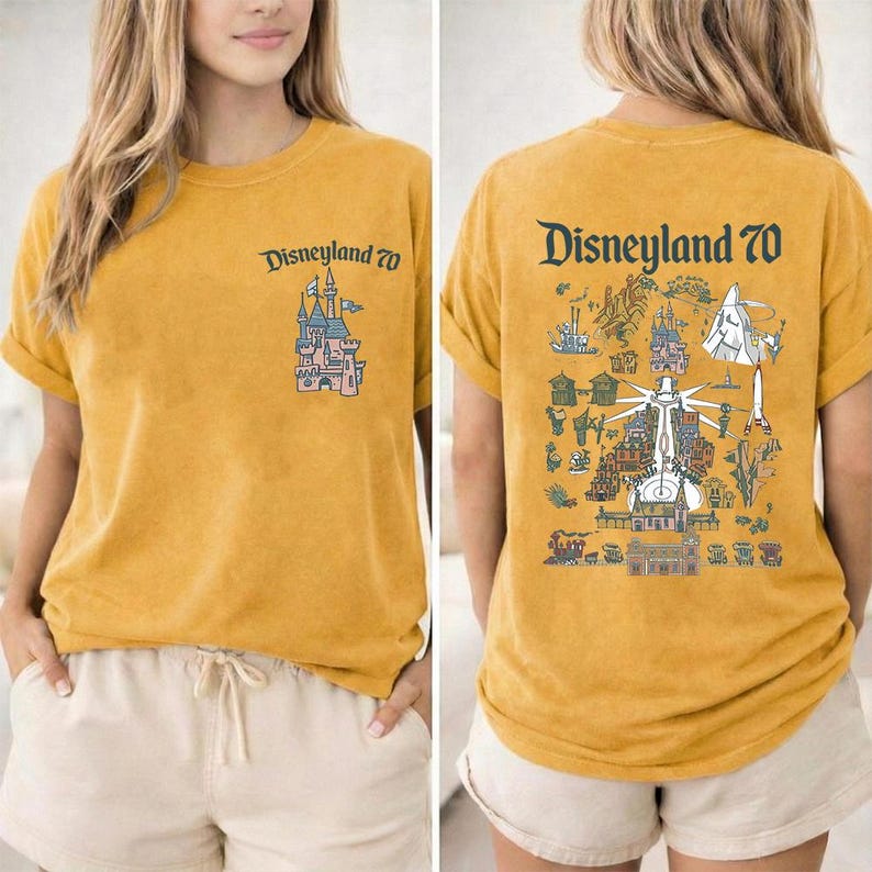Puede incluir: Una camiseta de color amarillo mostaza con un peque&ntilde;o gr&aacute;fico de un castillo y el texto Disneyland 70 en el pecho izquierdo. La parte trasera muestra una gran ilustraci&oacute;n detallada de varias atracciones y el texto Disneyland 70.