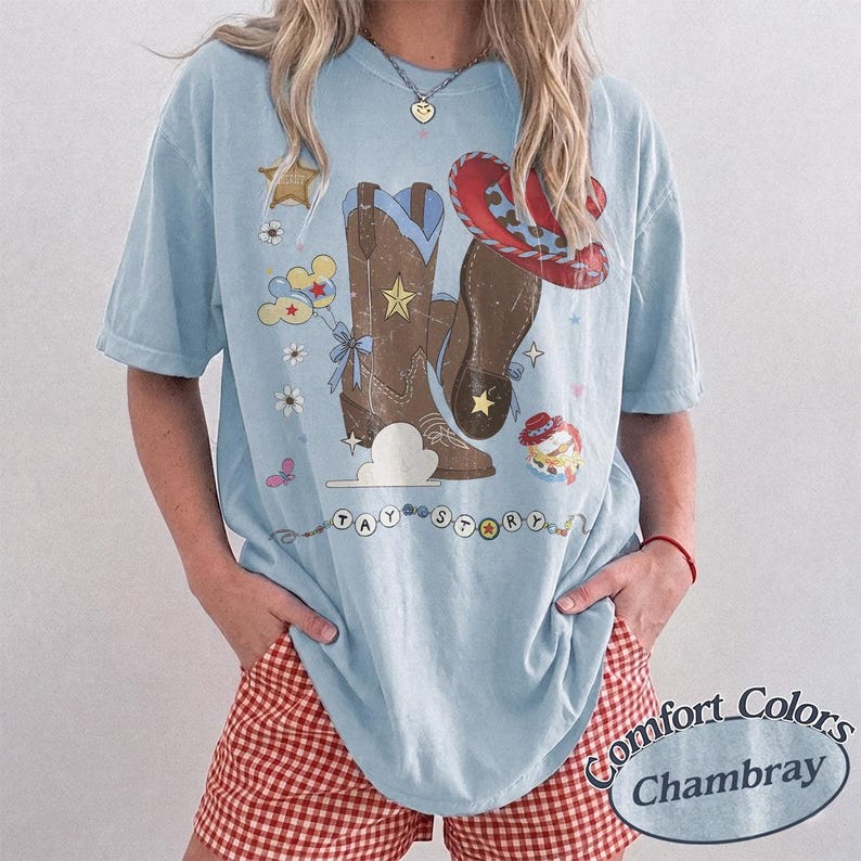 May include: A light blue short-sleeve t-shirt featuring a graphic print of brown cowboy boots, a red cowboy hat, a sheriff star, and clouds. The text TAY STORY is printed below the boots in a beaded style. The shirt has a crew neckline.
