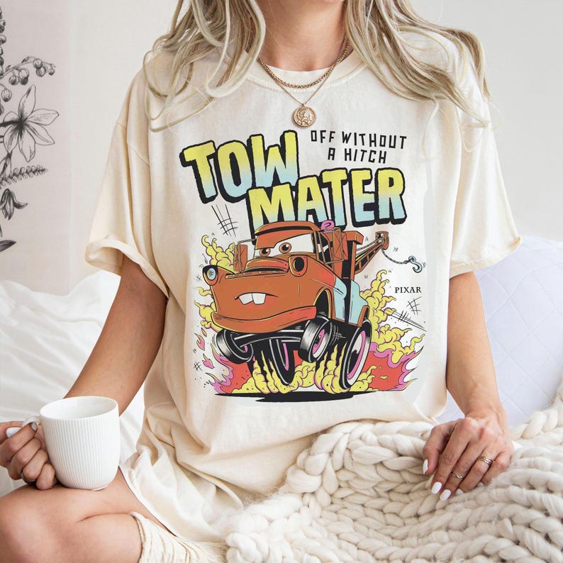 May include: Cream-coloured t-shirt with a cartoon image of Mater from the film Cars. The graphic includes the text "TOW MATER" and "OFF WITHOUT A HITCH" in a bold, colourful design. The image shows Mater with flames and the Pixar logo.