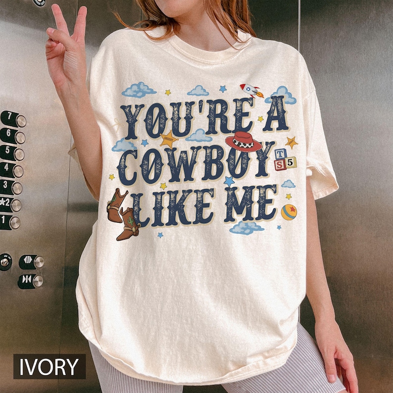 May include: An ivory short-sleeve t-shirt featuring the dark blue text "YOU'RE A COWBOY LIKE ME" surrounded by colourful graphics including a red cowboy hat, brown boots, a rocket ship, small clouds, yellow stars, a striped ball, and alphabet blocks.