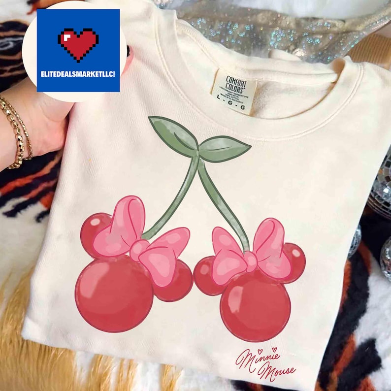 Op de afbeelding: Cr&egrave;mekleurige sweatshirt met een Minnie Mouse kersendesign. De afbeelding toont twee kersen met roze strikken en groene stelen. De woorden "Minnie Mouse" staan eronder in cursieve letters.