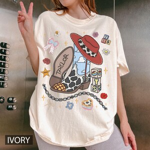 Puede incluir: Una camiseta color marfil con un estampado gr&aacute;fico de una bota vaquera con patr&oacute;n de vaca, un sombrero rojo, bloques de letras con la T, S y 5, un collar de cuentas con el texto I KNEW IT y el nombre TAYLOR en la suela de la bota.