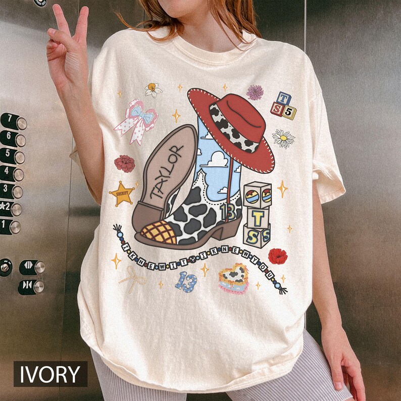 Puede incluir: Una camiseta color marfil con un estampado gr&aacute;fico de una bota vaquera con patr&oacute;n de vaca, un sombrero rojo, bloques de letras con la T, S y 5, un collar de cuentas con el texto I KNEW IT y el nombre TAYLOR en la suela de la bota.