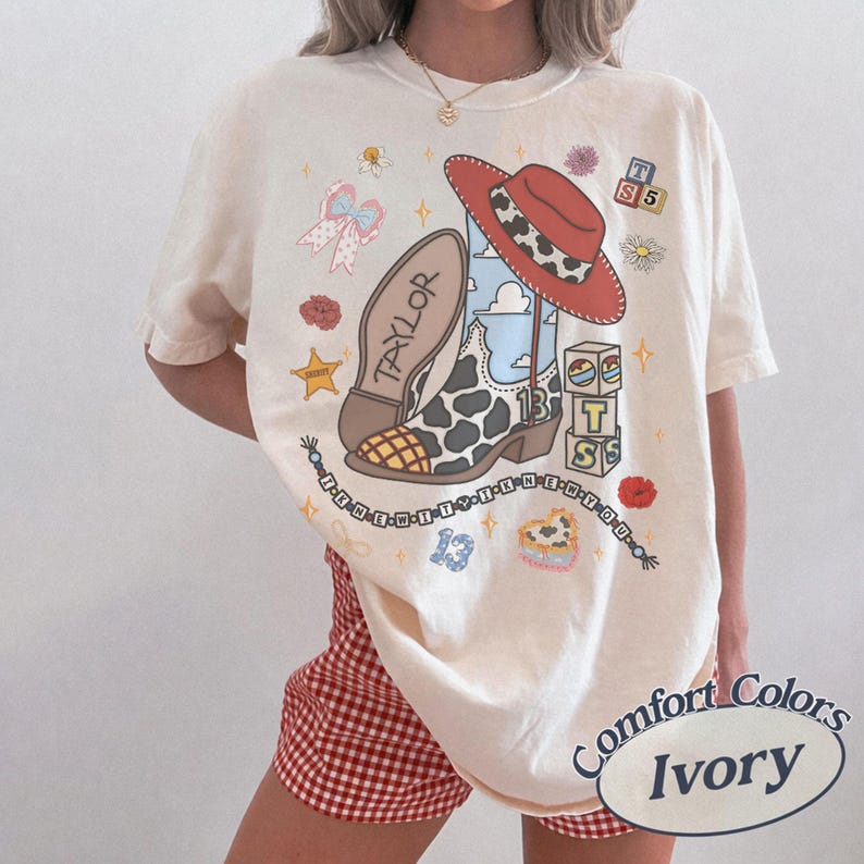 Puede incluir: Una camiseta color marfil con un estampado gr&aacute;fico de una bota de vaquero con patr&oacute;n de manchas de vaca, un sombrero de vaquero rojo y el nombre Taylor. El dise&ntilde;o incluye bloques de letras, un n&uacute;mero 13 azul, una cadena de cuentas y flores.