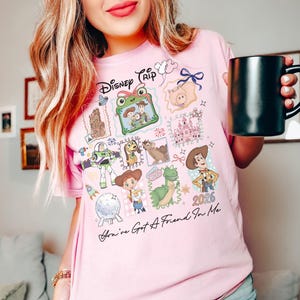 May include: A light pink t-shirt featuring a graphic print with the text Disney Trip and You've Got A Friend In Me. The design includes illustrations of Toy Story characters, a castle, and the year 2026, arranged in a collage of framed squares.