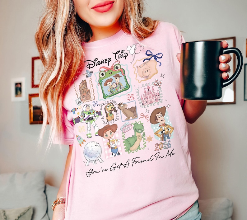 May include: A light pink t-shirt featuring a graphic print with the text Disney Trip and You've Got A Friend In Me. The design includes illustrations of Toy Story characters, a castle, and the year 2026, arranged in a collage of framed squares.
