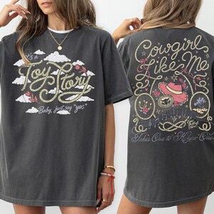 May include: A charcoal gray t-shirt featuring a front graphic with white clouds and the text It's a Toy Story Baby, just say yes. The back graphic displays a pink cowboy hat, rope accents, and the text Cowgirl like me Takes One to Know One.