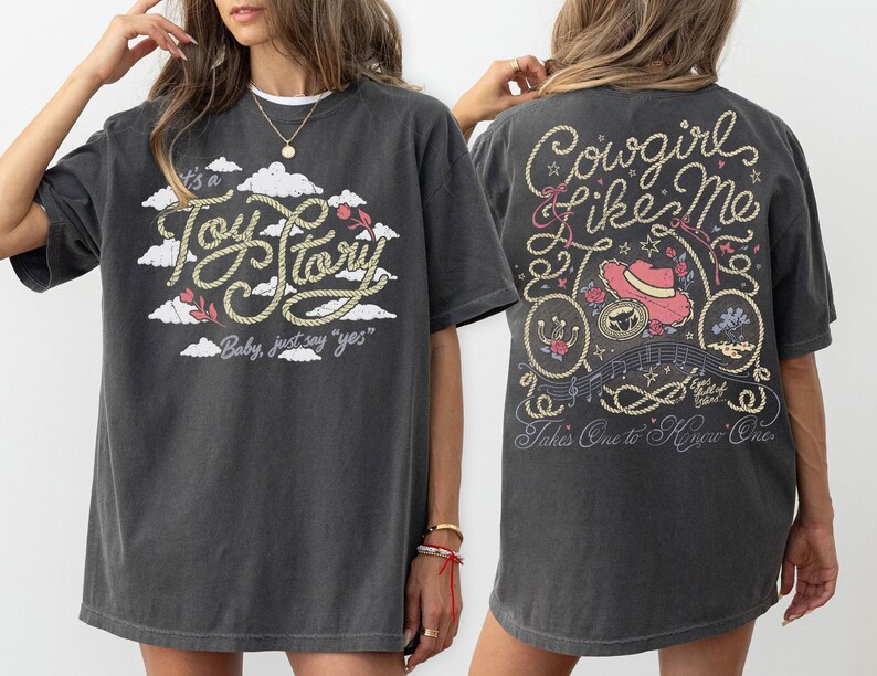 May include: A charcoal gray t-shirt featuring a front graphic with white clouds and the text It's a Toy Story Baby, just say yes. The back graphic displays a pink cowboy hat, rope accents, and the text Cowgirl like me Takes One to Know One.