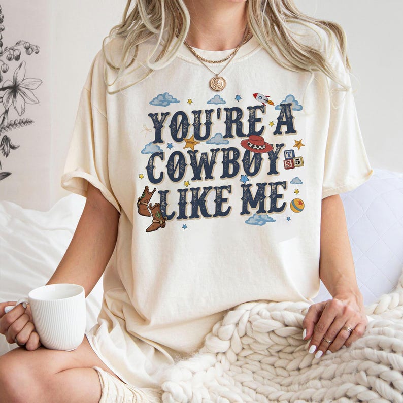 May include: A cream-coloured t-shirt featuring the dark blue text "YOU'RE A COWBOY LIKE ME" surrounded by illustrations of a red cowboy hat, brown boots, a rocket, stars, clouds, a colourful ball, and small blocks with the letters T, S, and 5.