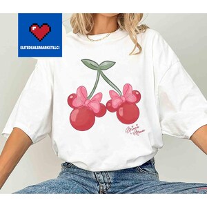 Op de afbeelding: Wit T-shirt met een cartoon kers ontwerp. De kersen zijn rood met roze strikken en groene stelen. De tekst "Minnie Mouse" is cursief onder de kersen geschreven. Het logo "ELITEDEALSMARKETLLC!" staat in de linkerbovenhoek.