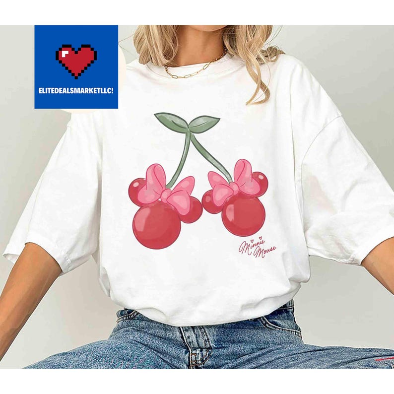 Op de afbeelding: Wit T-shirt met een cartoon kers ontwerp. De kersen zijn rood met roze strikken en groene stelen. De tekst "Minnie Mouse" is cursief onder de kersen geschreven. Het logo "ELITEDEALSMARKETLLC!" staat in de linkerbovenhoek.