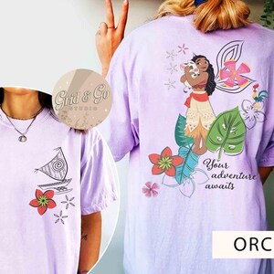 Może przedstawiać: Koszulki w kolorze orchidei z grafiką. Jedna koszulka przedstawia żagl&oacute;wkę i kwiaty, druga postać z kresk&oacute;wki z świnią, kwiatami i napisem "Your adventure awaits".