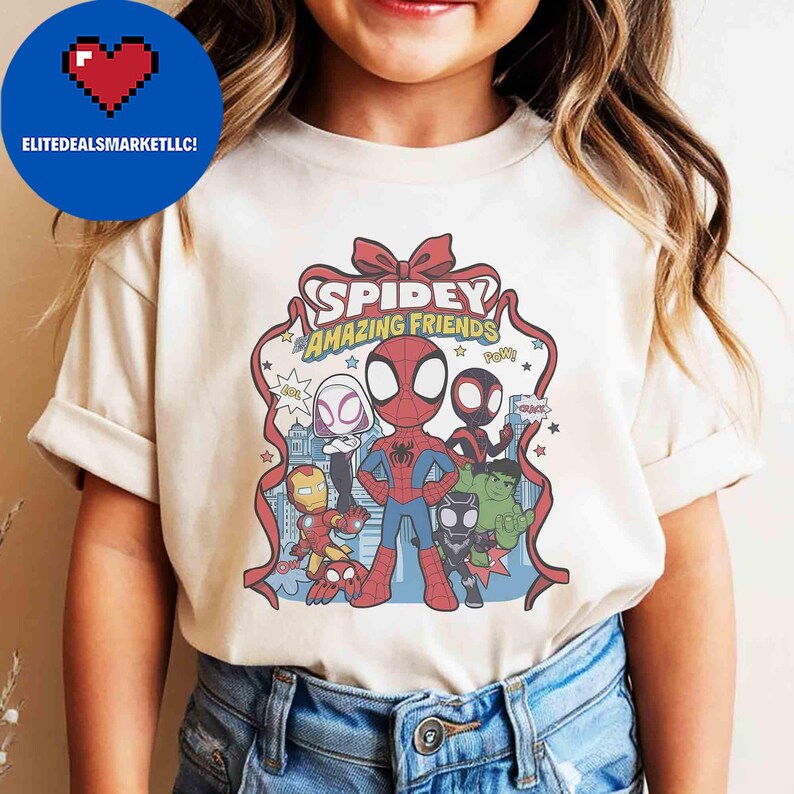 May include: A cream-colored t-shirt featuring a Spidey and His Amazing Friends graphic. The design includes Spiderman, Iron Man, Hulk, and other characters. The text "Spidey Amazing Friends" is displayed in a red ribbon design.