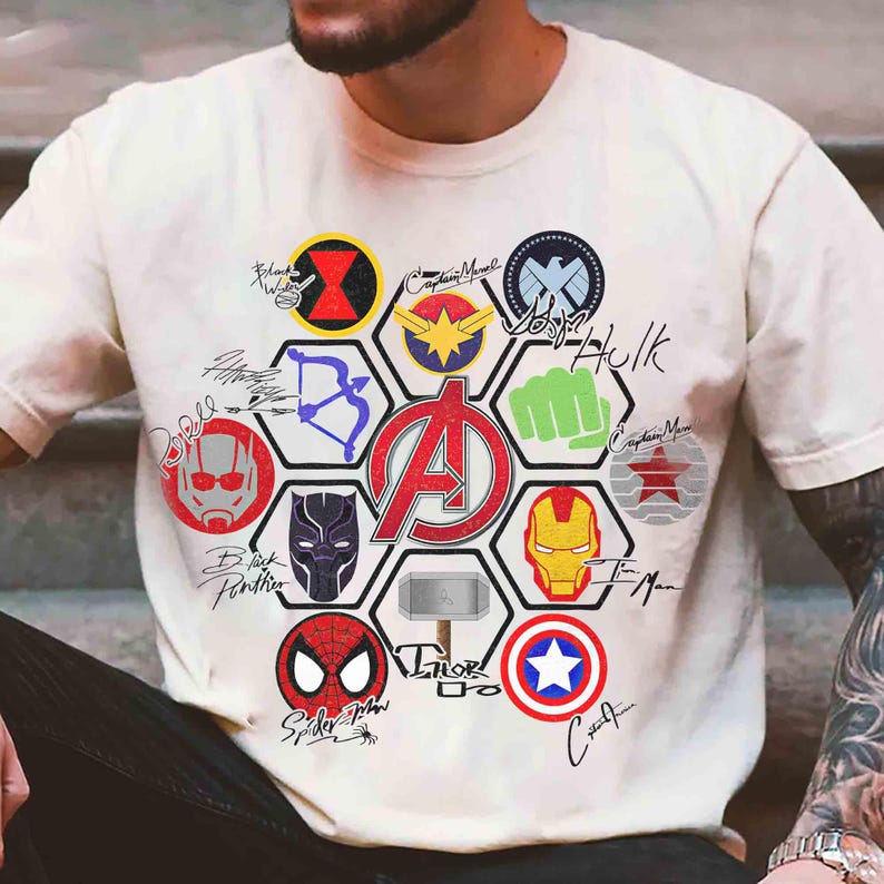Pu&ograve; includere: T-shirt color crema con un grande logo degli Avengers al centro, circondato da vari emblemi e firme di supereroi. Include Iron Man, Captain Marvel, Spider-Man e Thor. Il design &egrave; una grafica colorata.