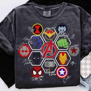 Pu&ograve; includere: T-shirt grigio scuro con loghi e firme di supereroi colorati. Il design include il logo degli Avengers al centro, circondato da emblemi di personaggi e firme. La maglietta &egrave; realizzata in un materiale morbido e confortevole.