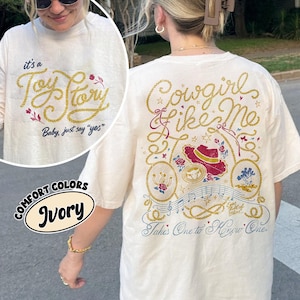 May include: An ivory t-shirt featuring a graphic design on the back with the text Cowgirl Like Me, Eyes full of stars, and Takes One to Know One. The design includes a red hat, musical notes, and rope motifs. The front shows It's a Toy Story Baby, just say yes.